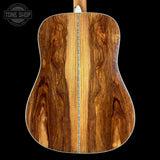 The back of an acoustic guitar made of rich, curly brown wood, featuring a central soundhole and a white edge trim, shown against a solid black background with a Tone Shop watermark in the top left.