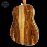 The back of an acoustic guitar made of rich, dark brown wood with prominent grain patterns and a light-colored binding, shown against a solid black background with a Tone Shop watermark in the top left.