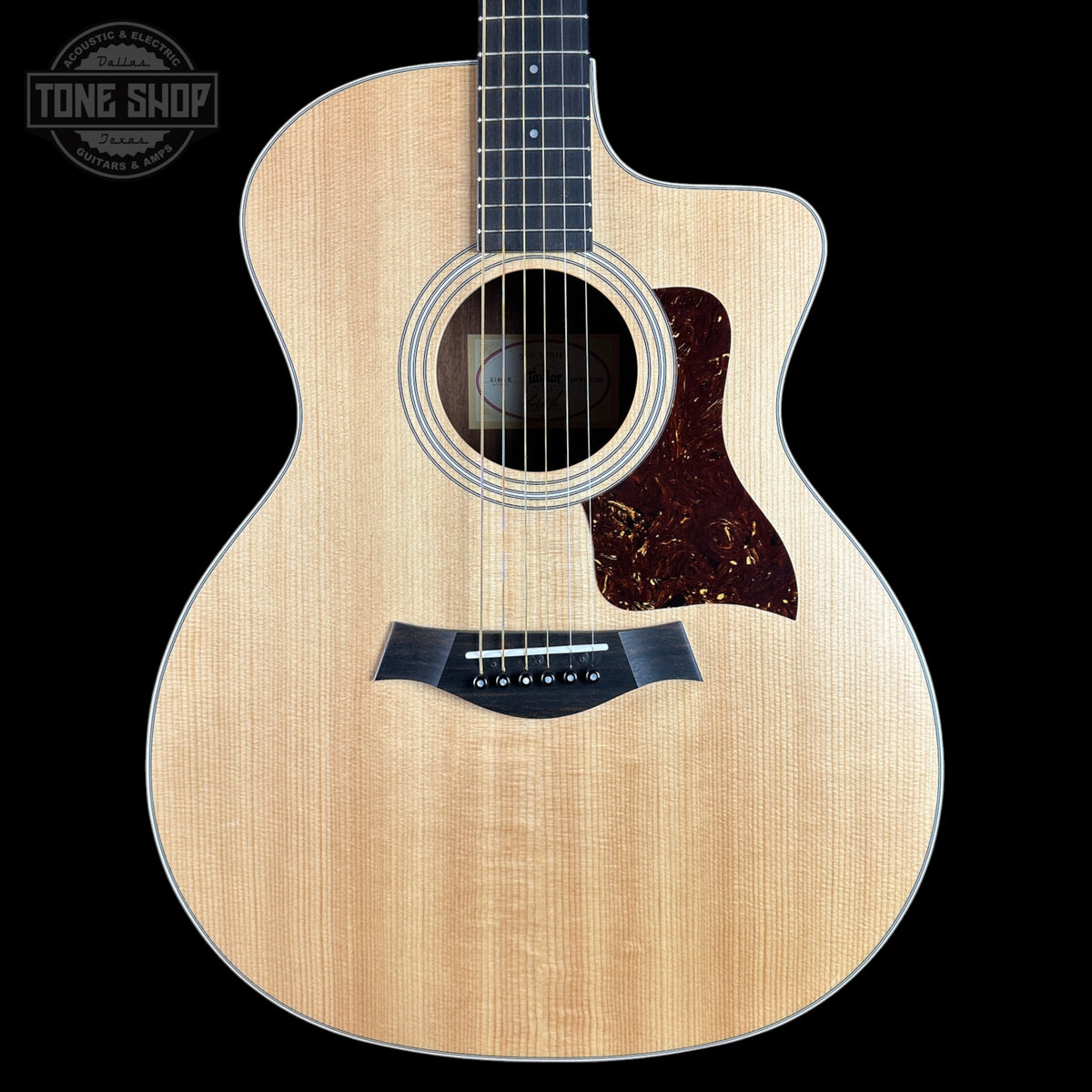 A Taylor acoustic guitar with a natural wood finish and a dark tortoiseshell pickguard, shown against a solid black background with a Tone Shop watermark in the top left corner.