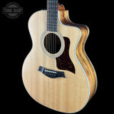 A Taylor acoustic-electric guitar with a natural wood body, dark pickguard, and cutaway side, shown against a solid black background with a Tone Shop watermark in the top left corner.