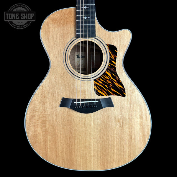 A Taylor acoustic-electric guitar with a natural wood finish and a black-and-orange tiger-eye style pickguard, shown against a solid black background with a Tone Shop watermark in the top-left corner.