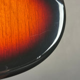 Close-up of a glossy sunburst electric guitar body edge, showing the warm orange-to-brown wood finish fading into black, with a light gray background.