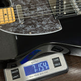 A close-up of an electric guitar body resting on a digital scale, showing the scale's display reading 7.154 lbs and its buttons, with the guitar's black pickguard and wood neck visible.