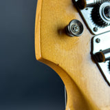 Close-up detail of a light natural wood electric guitar body with a metal tuning peg and gear mechanism against a dark gray background.