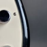 Close-up detail of an electric guitar body showing a cream-colored pickguard, a black control knob, a black edge trim, and a silver screw against a dark gray background.