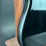 Close-up of a glossy black curved object, likely part of a guitar or similar instrument, standing beside a wooden stand against a dark gray background.
