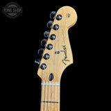 Close-up of the headstock of a Fender Stratocaster electric guitar, showing the natural maple wood finish, six black tuning pegs, and the Fender logo, set against a solid black background.