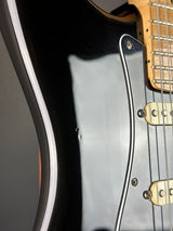 Close-up of an electric guitar body showing a glossy black finish, a white pickguard, cream-colored hardware, and part of the light wood neck.