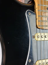 Close-up of a black electric guitar body with a white pickguard, gold hardware, and part of the natural wood neck.