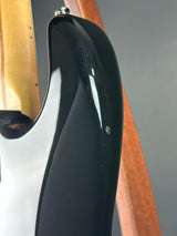 Close-up of a glossy black electric guitar body with a white edge, showing part of the wood neck and a brown wooden stand against a gray background.