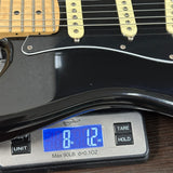 Close-up of a black electric guitar body resting on a digital scale showing 8 lbs 12 oz, with the guitar's cream pickups and maple neck visible.