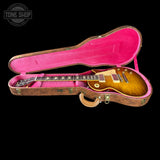 An electric guitar with a natural wood finish resting inside an open hard case with bright pink interior lining, shown against a solid black background. A Tone Shop watermark is visible in the top left corner.