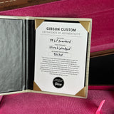 An open Gibson Custom presentation box showing a Certificate of Authenticity insert, with a pink velvet interior lining and a small pink bow visible at the bottom right.