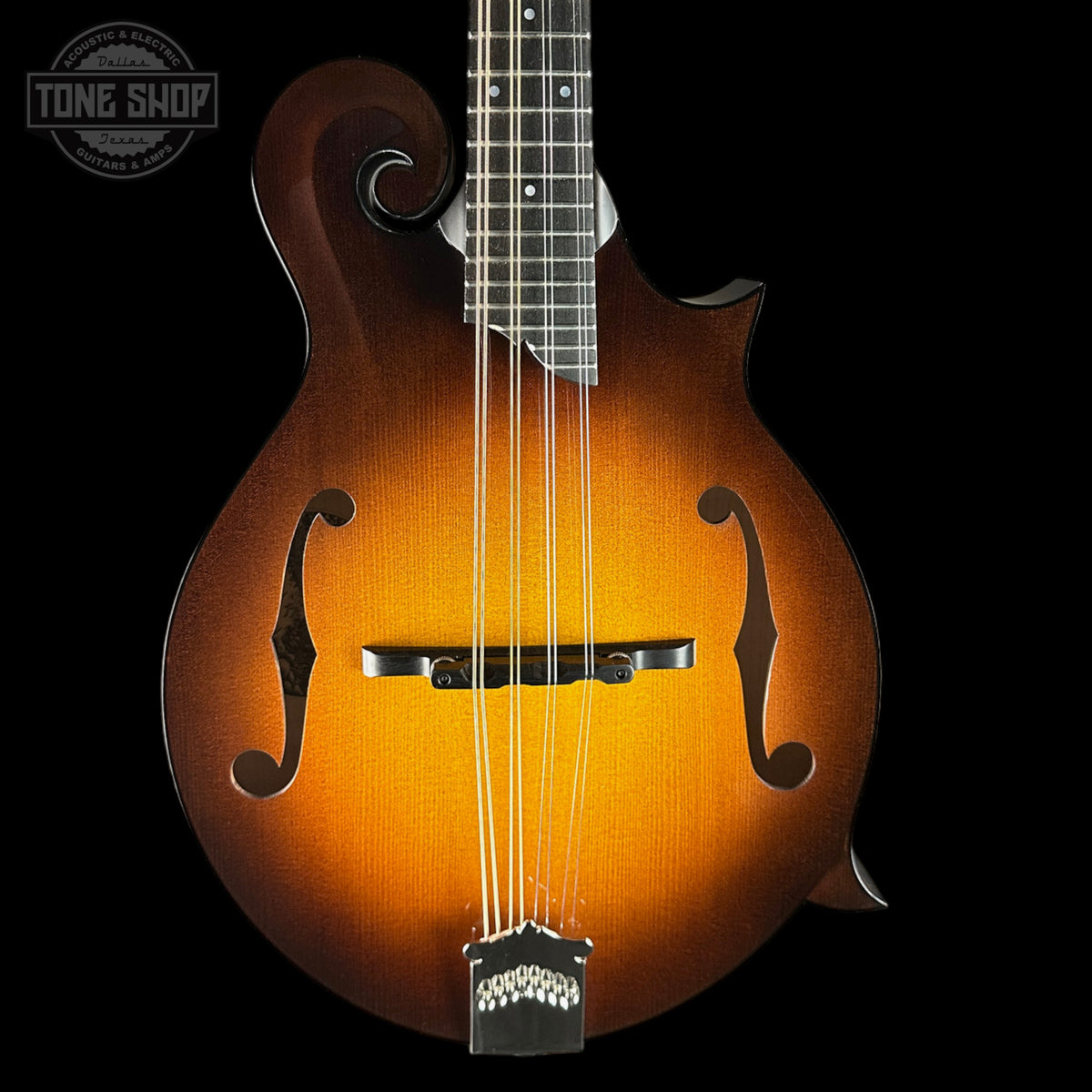 A mandolin with a classic sunburst finish, transitioning from a dark brown outer edge to a bright amber-orange center. The mandolin features a dark fretboard, silver metal tuning pegs, and a white chin bar, set against a solid black background with a Tone Shop watermark in the upper left corner.