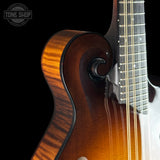 Close-up of a mandolin guitar body showing the rich amber-to-brown sunburst finish, scroll-shaped back, and part of the dark fretboard against a black background. A Tone Shop watermark is visible in the upper left.