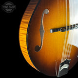 Close-up of a mandolin guitar body showing the sunburst finish, f-hole, bridge, and strings against a black background, with a Tone Shop watermark in the upper left.