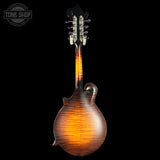 A mandolin shown from the back against a solid black background, featuring a glossy dark brown wood finish with a warm amber glow and a decorative scroll on the back body. A Tone Shop watermark is visible in the top-left corner.