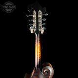 Close-up of the headstock and upper neck of a mandolin against a black background, with six silver tuning pegs and a glossy dark wood body showing warm amber reflections. A Tone Shop watermark appears in the upper left.