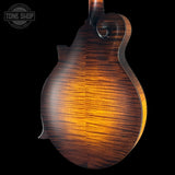 Close-up of the back of a mandolin with a rich, warm brown and amber wood grain finish, set against a solid black background with a Tone Shop watermark in the top-left corner.