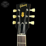 Close-up of a Gibson acoustic guitar headstock with six cream-colored tuning pegs, silver tuning keys, and a gold Gibson logo and arrow emblem on a black background.