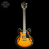 A classic electric guitar with a sunburst finish, featuring a black headstock, black pickguard, and gold-toned hardware, shown against a solid black background with a Tone Shop watermark in the top left corner.