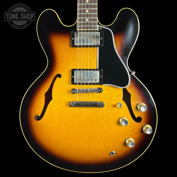 A classic electric guitar with a sunburst finish, featuring a bright yellow-orange center fading to dark brown and black edges. It has a black pickguard, chrome hardware, and gold-toned control knobs, shown against a solid black background with a Tone Shop watermark in the top left corner.
