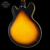 Close-up of the back of an electric guitar body showing a classic sunburst finish that transitions from bright amber-orange in the center to dark brown and black around the edges, with a thin gold-toned edge outline. A Tone Shop watermark is visible in the top left corner.