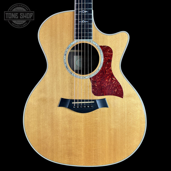 A Taylor acoustic-electric guitar with a natural wood finish and a dark red tortoiseshell pickguard, shown against a solid black background with a Tone Shop watermark in the top left corner.