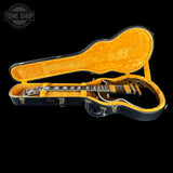 A black electric guitar with a glossy black finish and white binding, displayed inside an open black guitar case with a bright orange-yellow velvet interior. The guitar is positioned horizontally across the open case.