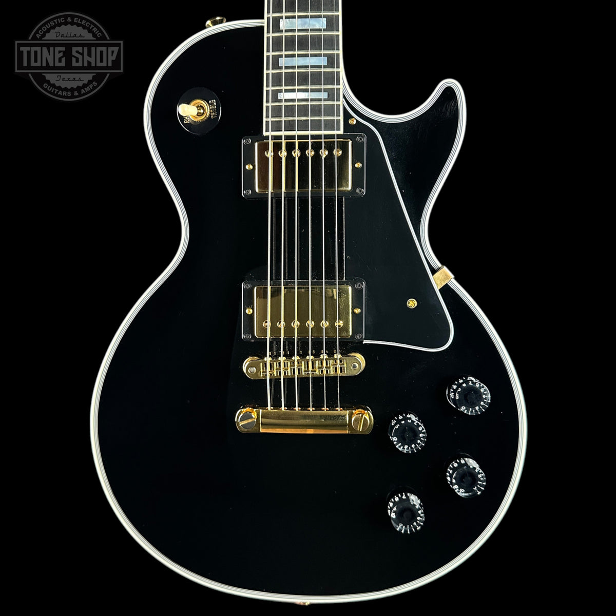 A black electric guitar with gold hardware and white binding, shown from a front-facing angle against a black background. A Tone Shop logo is visible in the top-left corner.