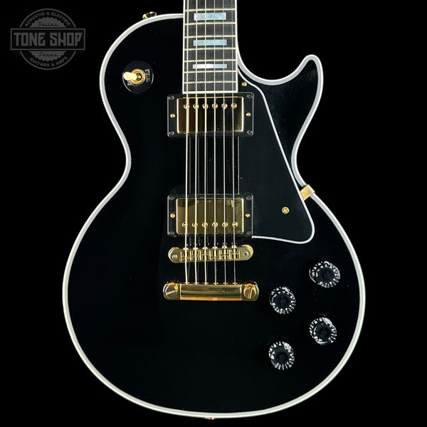A black electric guitar with gold hardware and white binding, shown from a front-facing angle against a black background. A Tone Shop logo is visible in the top-left corner.