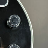Close-up detail of a black electric guitar body showing a volume control knob, a white binding edge, and part of another knob against a neutral gray background.