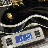 Close-up of a glossy black electric guitar body resting on a digital scale displaying 11.50 lbs, with gold hardware and part of the fretboard visible.