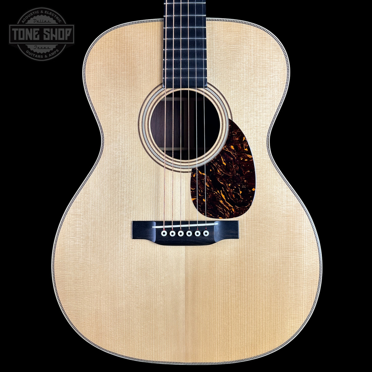 A front-facing acoustic guitar with a natural wood finish and dark pickguard, shown against a solid black background with a Tone Shop watermark in the top-left corner.