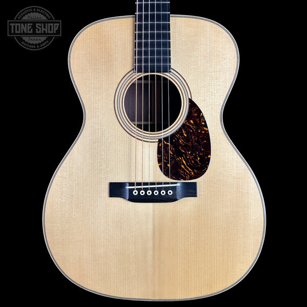 A front-facing acoustic guitar with a natural wood finish and dark pickguard, shown against a solid black background with a Tone Shop watermark in the top-left corner.