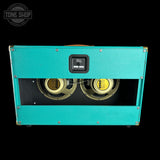 A turquoise teal guitar speaker cabinet shown from the front against a solid black background. The cabinet features a textured teal finish, a central black control panel, and a lower glass-fronted section revealing two gold and black speaker cones.