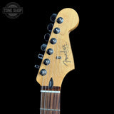 Close-up of a Fender Stratocaster electric guitar headstock in natural maple wood, featuring six black tuning pegs and the Fender logo, set against a solid black background.