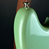 Close-up of a glossy light green guitar body with a silver metal tuning peg visible at the top, photographed against a dark background.