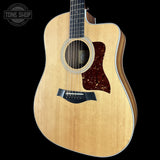 A Taylor acoustic-electric guitar with a natural wood finish and a dark tortoiseshell pickguard, shown against a solid black background with a Tone Shop watermark in the top-left corner.