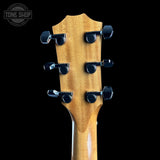 Close-up of the headstock of a guitar with a natural wood finish and six black tuning pegs, shown against a solid black background. A Tone Shop watermark is visible in the top left corner.