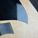 Close-up of a guitar body showing a black pickguard and a brushed metal or dark gray hardware piece on a light natural wood grain surface.