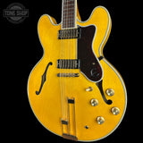 A close-up of a yellow-gold double-cutaway electric guitar against a black background, showing the body, pickups, f-holes, and control knobs. A Tone Shop watermark is visible in the top-left corner.
