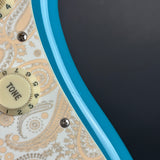 Close-up of an electric guitar body showing a bright turquoise blue edge, a cream tone control dial, and a white pickguard with a beige paisley pattern against a dark gray background.