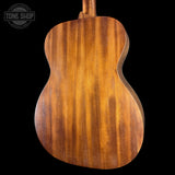 Close-up of the back of an acoustic guitar showing a rich, warm brown wood grain finish against a solid black background, with a Tone Shop watermark in the top left corner.