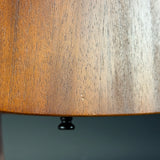 Close-up of a wooden lampshade with visible wood grain and a small black finial at the bottom, photographed against a dark blue-gray background.