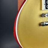 Close-up of the lower bout corner of an electric guitar with a metallic gold finish, a red edge stripe, a white binding, and chrome hardware.