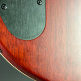 Close-up detail of an electric guitar body showing a reddish-brown wood grain finish, a dark brushed metal pickguard, and part of a silver hardware edge.