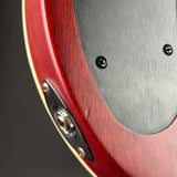 Close-up of a red electric guitar body showing the wood grain finish, a brushed silver metal pickguard, and a silver hardware plate with a screw-in jack.