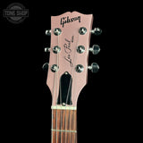 Close-up of a Gibson Les Paul Model acoustic guitar headstock in a metallic rose gold finish, featuring six silver tuning pegs and a black soundhole cover, set against a solid black background.
