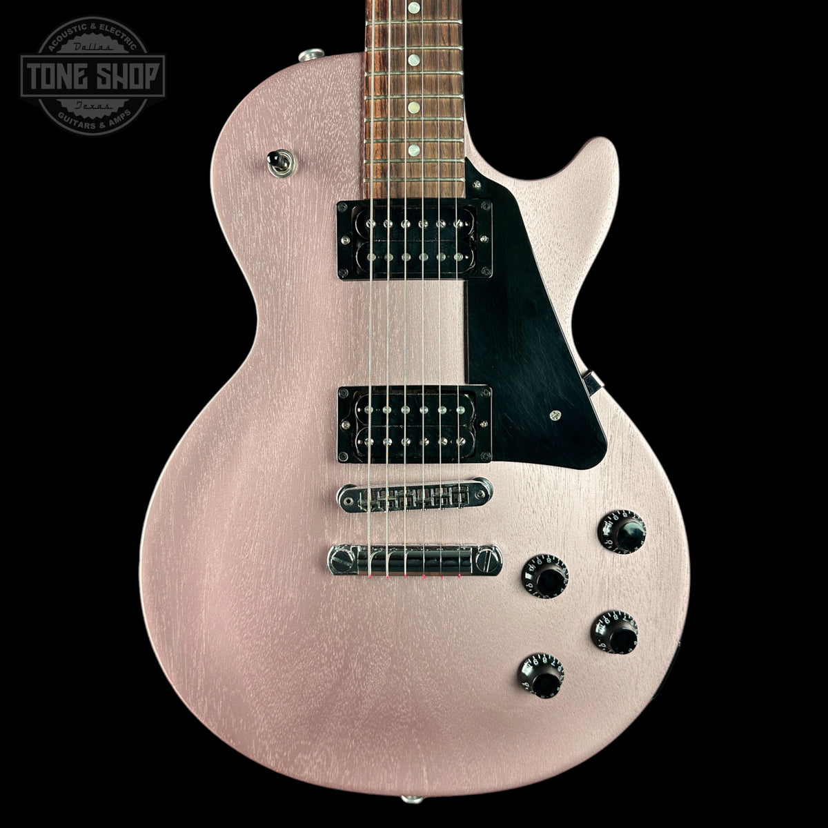 A close-up of an electric guitar body in a metallic rose gold finish with a black pickguard and black hardware, shown against a solid black background. A Tone Shop watermark is visible in the top-left corner.